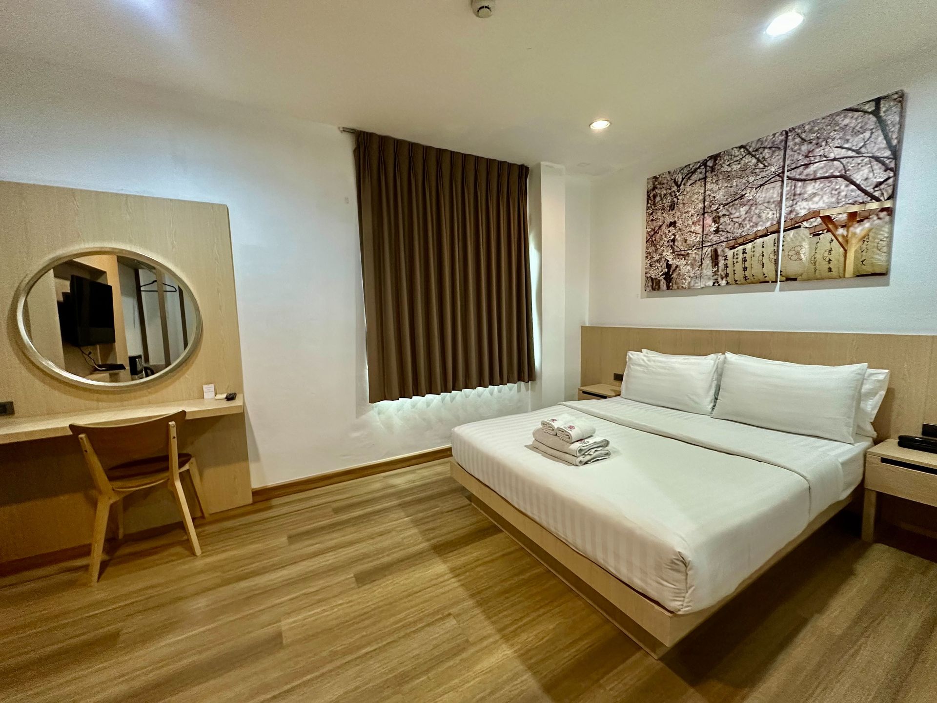 Hotel room with a bed, vanity, and window with brown curtains. A landscape picture hangs above the bed.