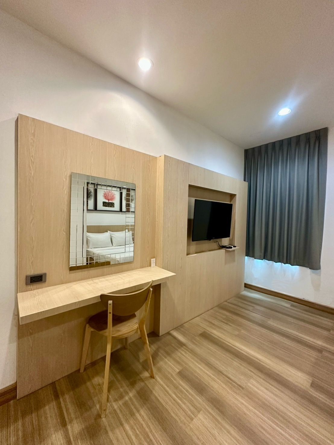 Hotel room with wooden desk, mirror, TV, and window with gray curtains.