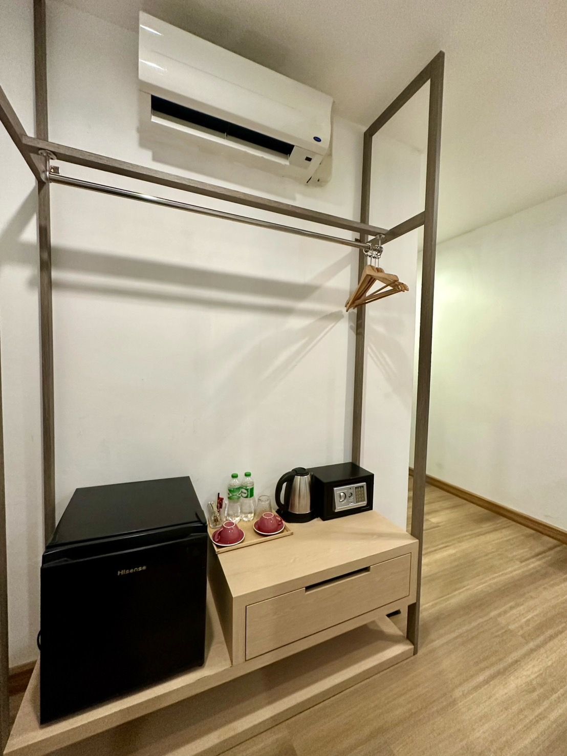 A room's built-in closet with a mini-fridge, safe, electric kettle, and water bottles. Air conditioner mounted above.