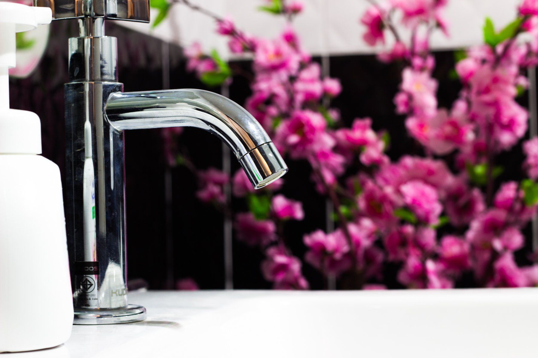 Chrome faucet on white sink, soap dispenser left, pink flowers background.