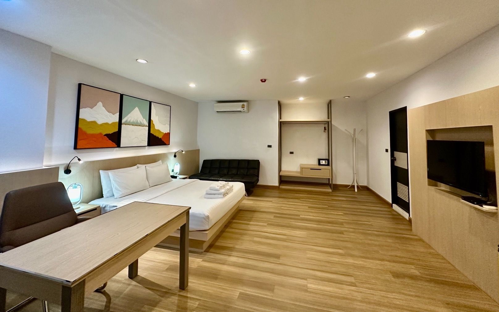 Hotel room with bed, desk, sofa, and artwork on the wall. Light wood flooring and neutral tones.