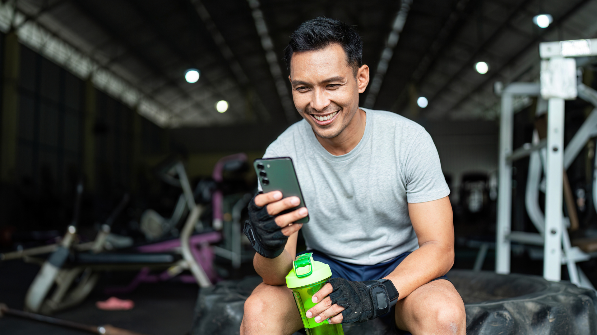 Man in gym, smiling at phone. Holding water bottle, sitting on tire. Dark gym in background.