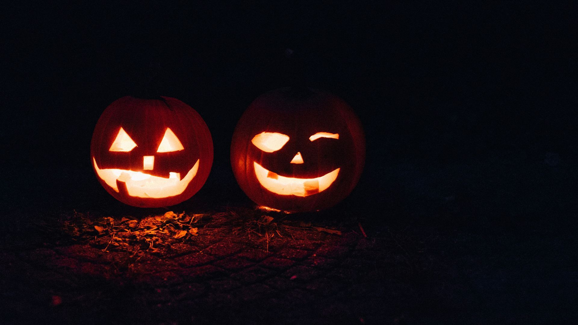 Two lit jack-o'-lanterns with carved faces glow against a dark background, one winking.