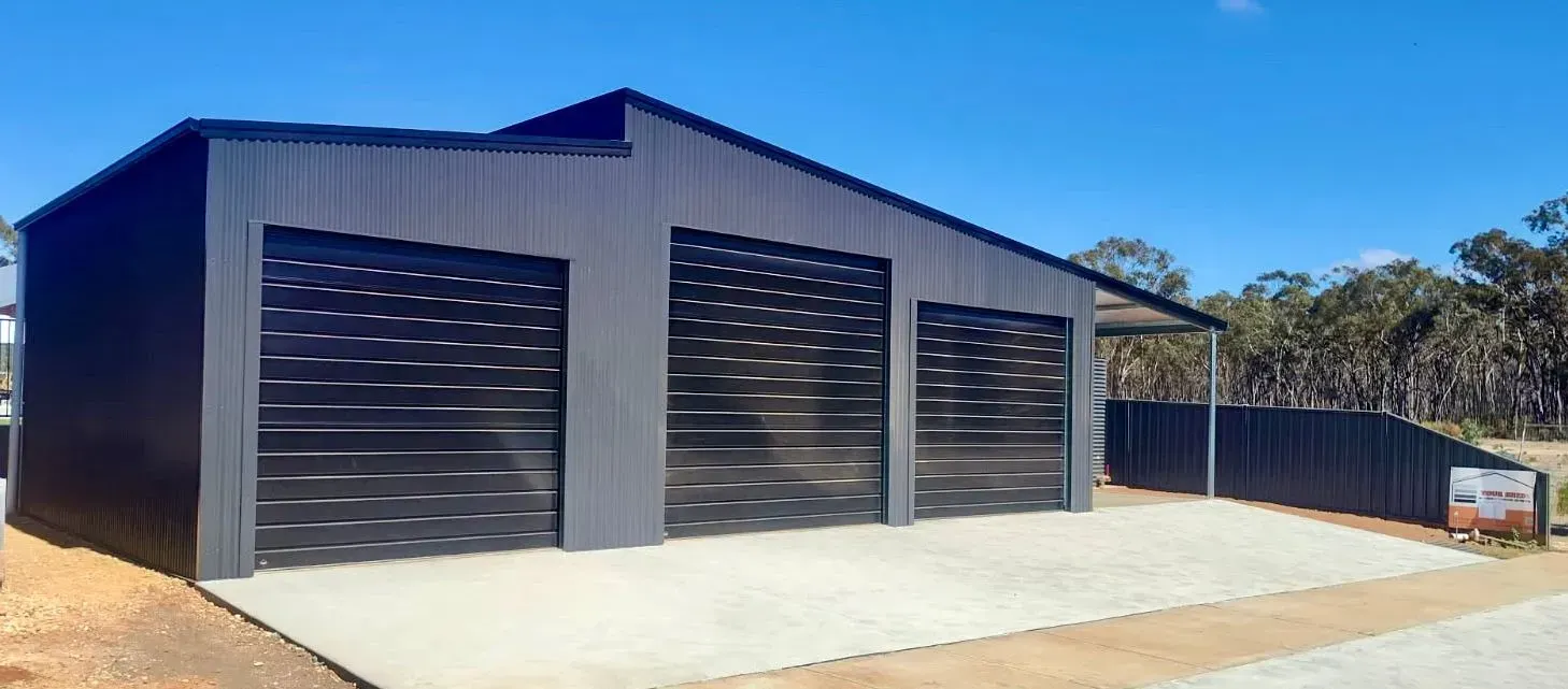 Modern gray metal shed with three black roller doors and a concrete apron under a blue sky
