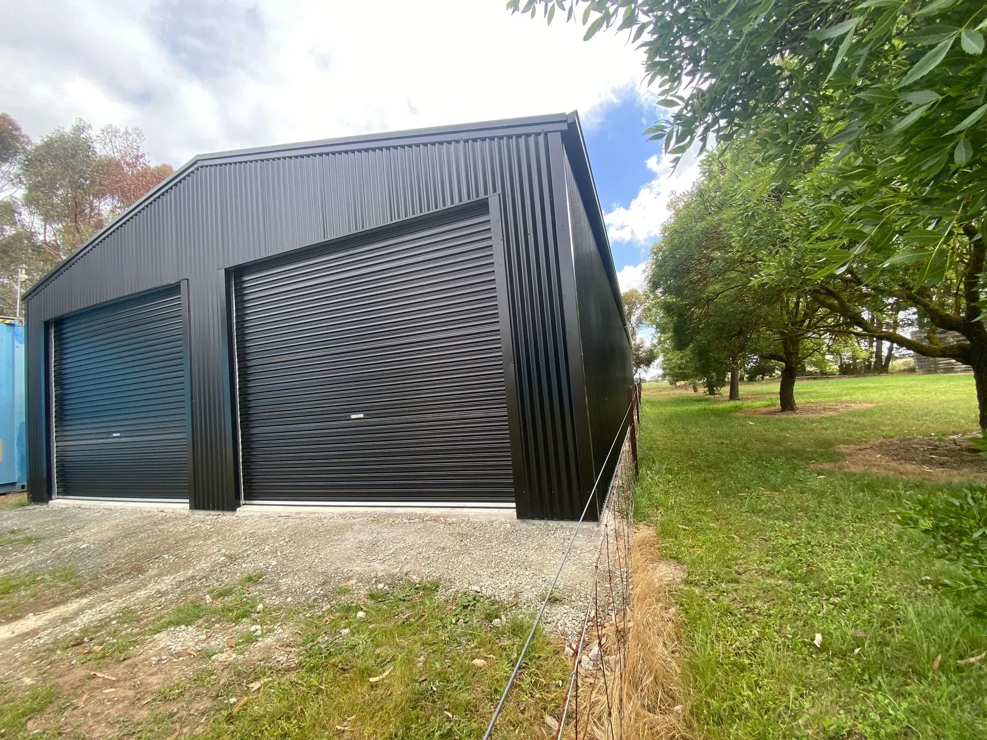 Black metal shed with two roller doors on a gravel pad beside grassy trees under a cloudy sky