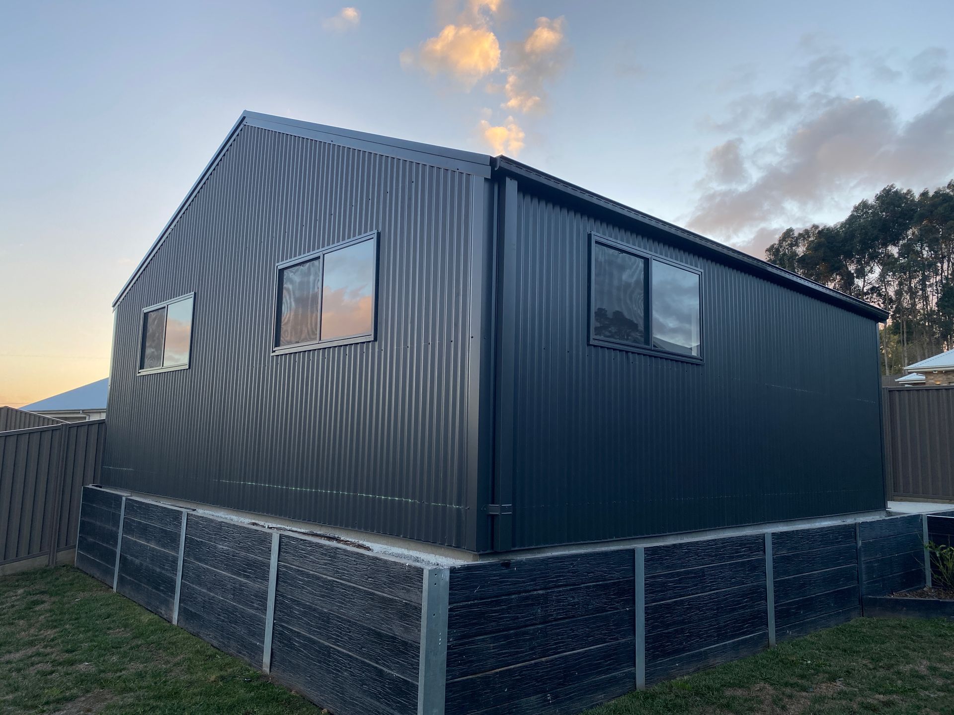 Dark gray modular house exterior at dusk with small windows and a fenced base.