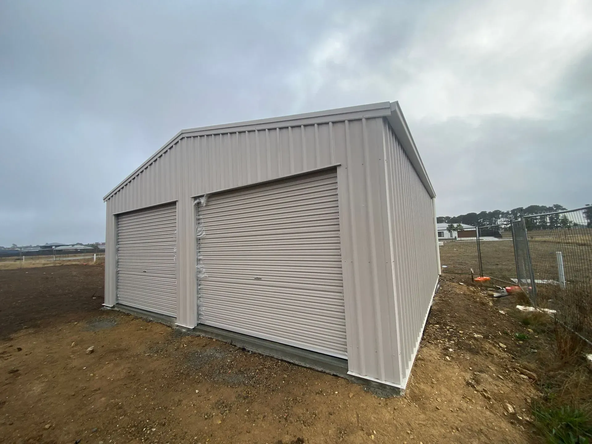 Small gray metal shed with roll-up doors on a dirt lot under a cloudy sky