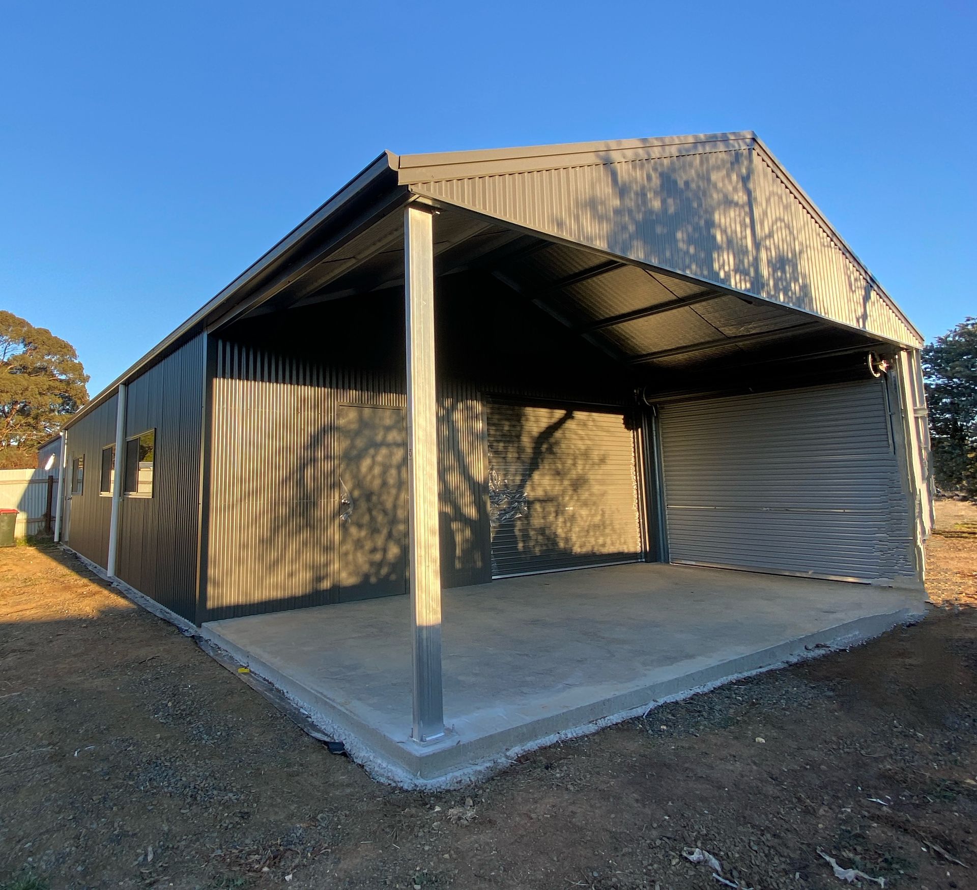 Small corrugated-metal shed with open front, concrete pad, and roller door in a sunny yard