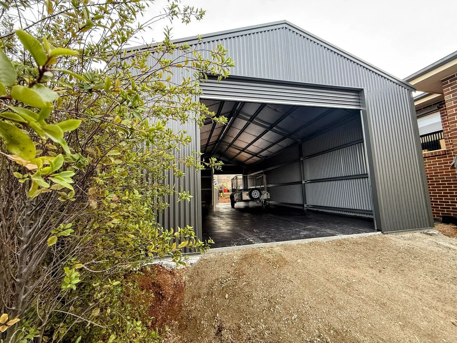 Open-front gray metal shed with a tarp inside, beside bushes and a gravel slope.
