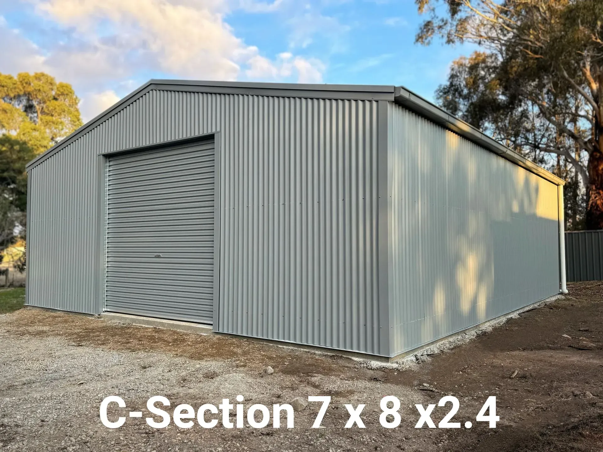 Gray steel storage shed with roll-up door, labeled “C-Section 7 x 8 x2.4,” outdoors under trees