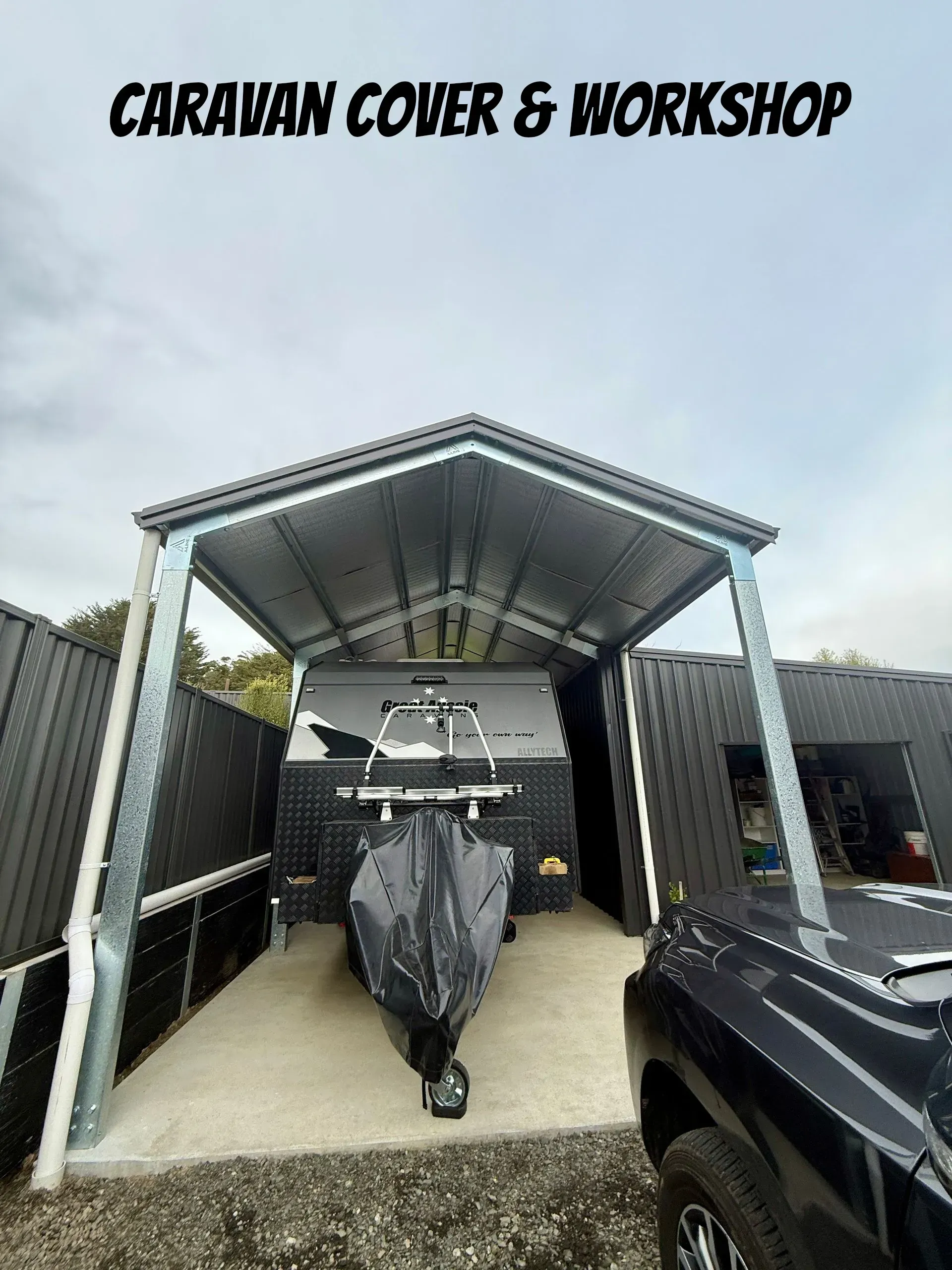 Covered caravan workshop with a boat trailer centered under a metal roof, black vehicle at right.