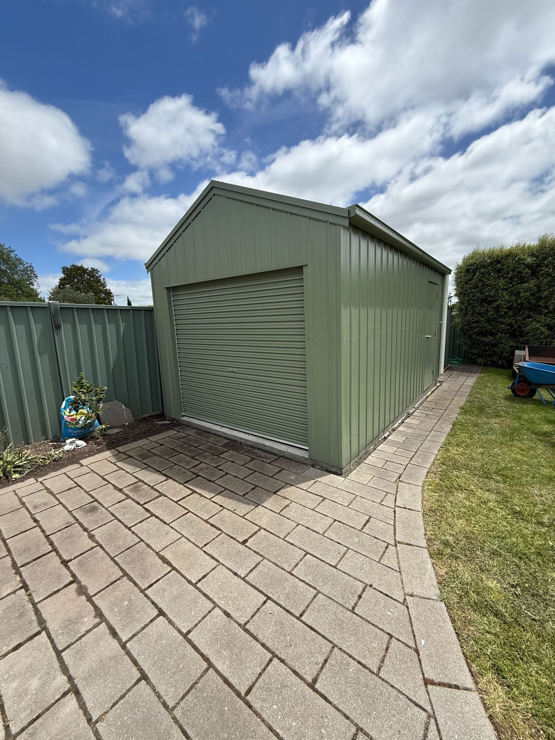 Green garden shed with a closed roll-up door on a paved patio under a blue sky