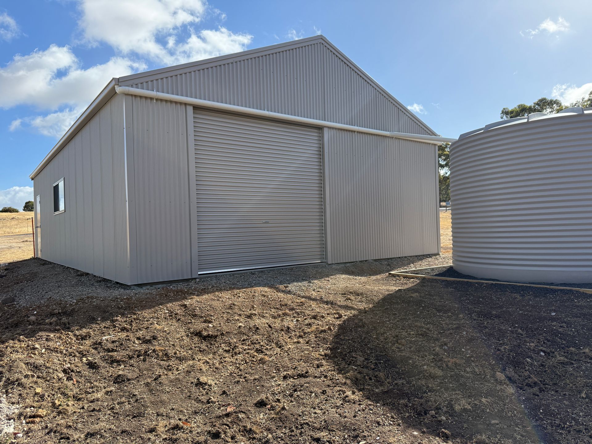 Gray metal shed with a closed roll-up door beside a large water tank on a dirt lot under a blue sky