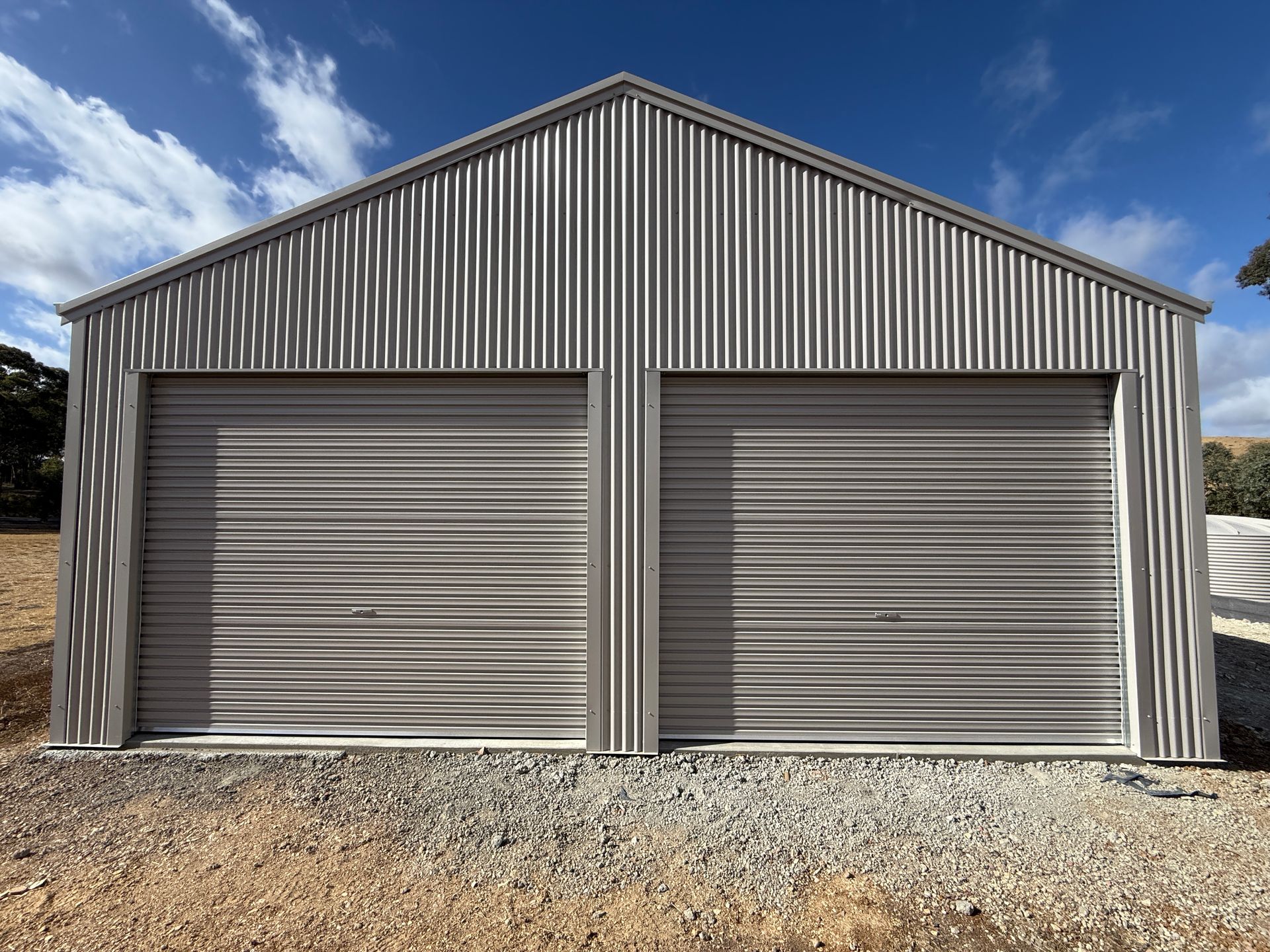 Two-door metal garage with gray corrugated siding and roll-up doors on a gravel lot under a blue sky