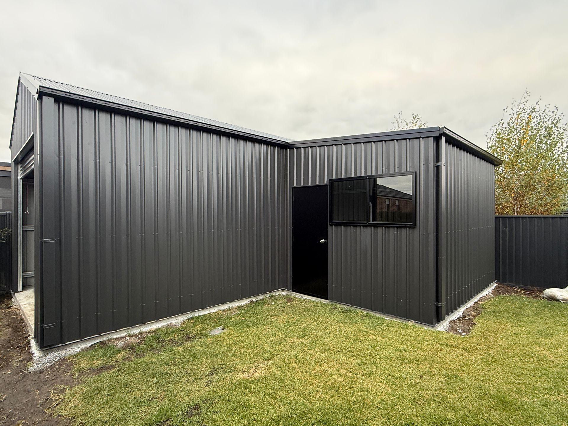 Black corrugated metal shed with a small window, set on a grassy yard under a cloudy sky.