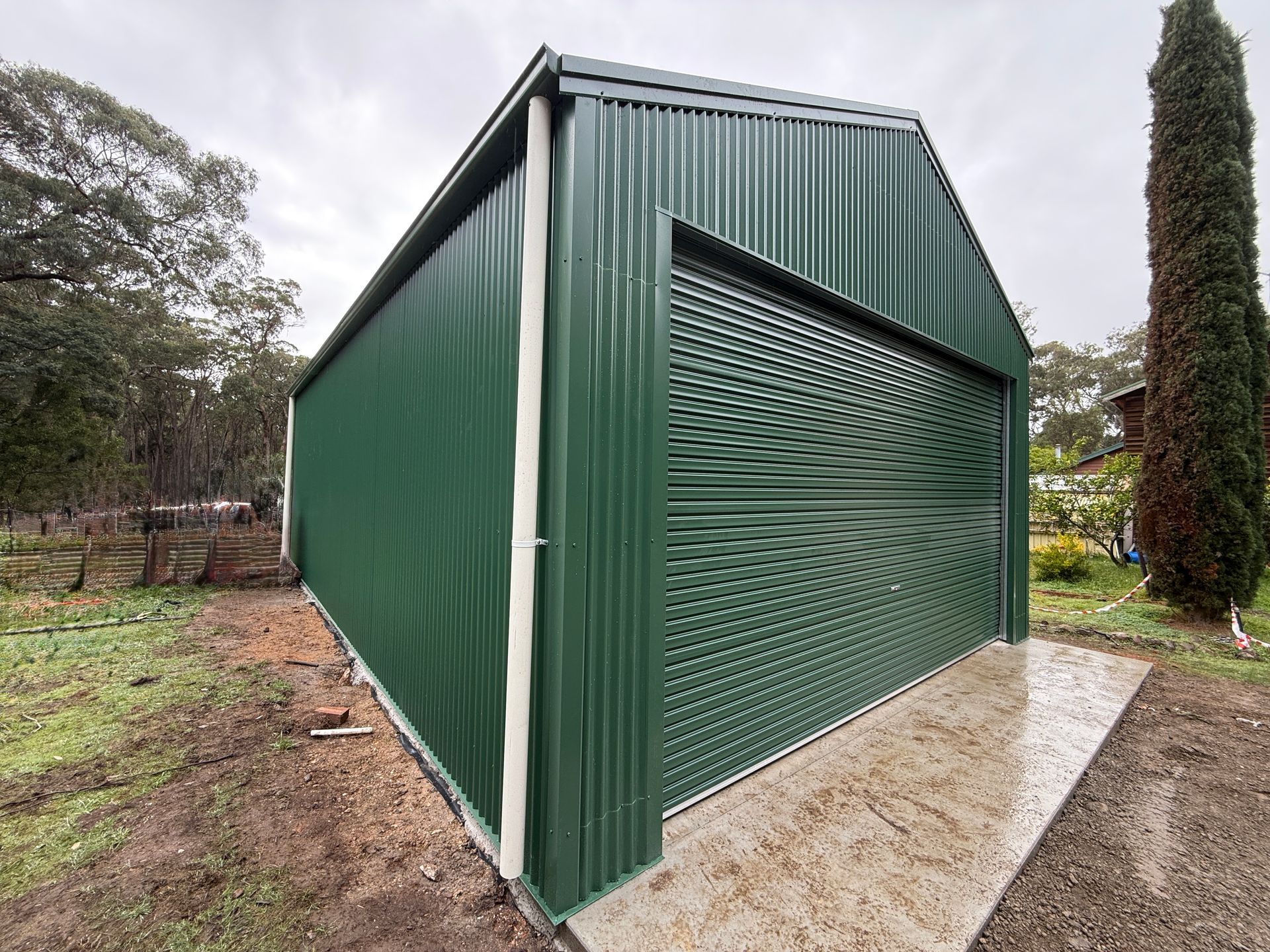 Green corrugated metal shed with a sloped roof on a concrete pad, surrounded by trees and dirt.