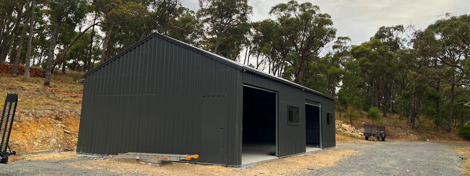 Dark gray metal shed with open doors on a dirt lot, surrounded by trees and a rocky hillside.