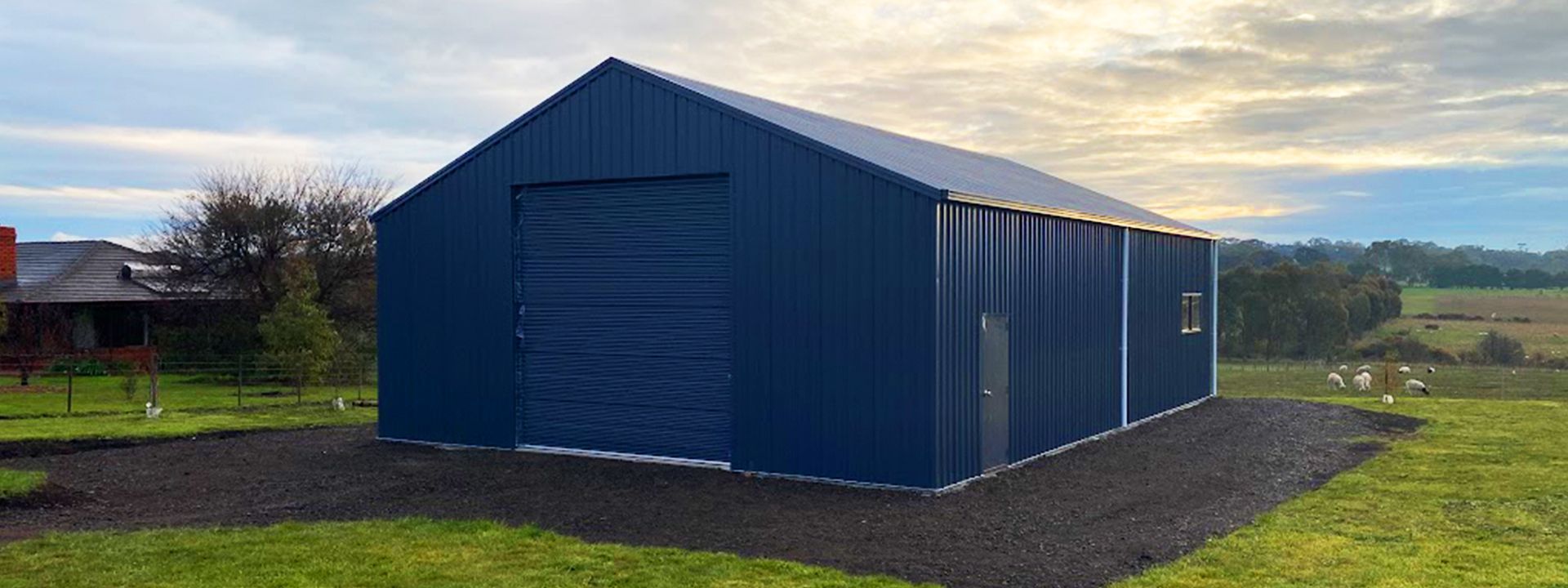 Blue metal shed or garage in a grassy field at sunset, with a large roller door and open countryside background