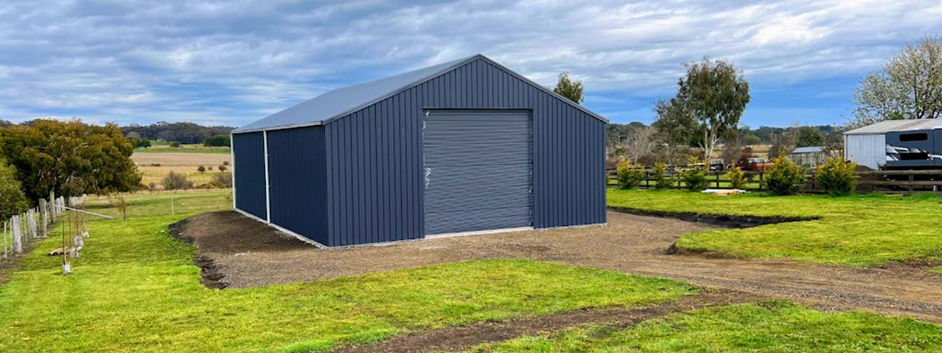 Blue metal shed with a roller door in a grassy rural field under a cloudy sky