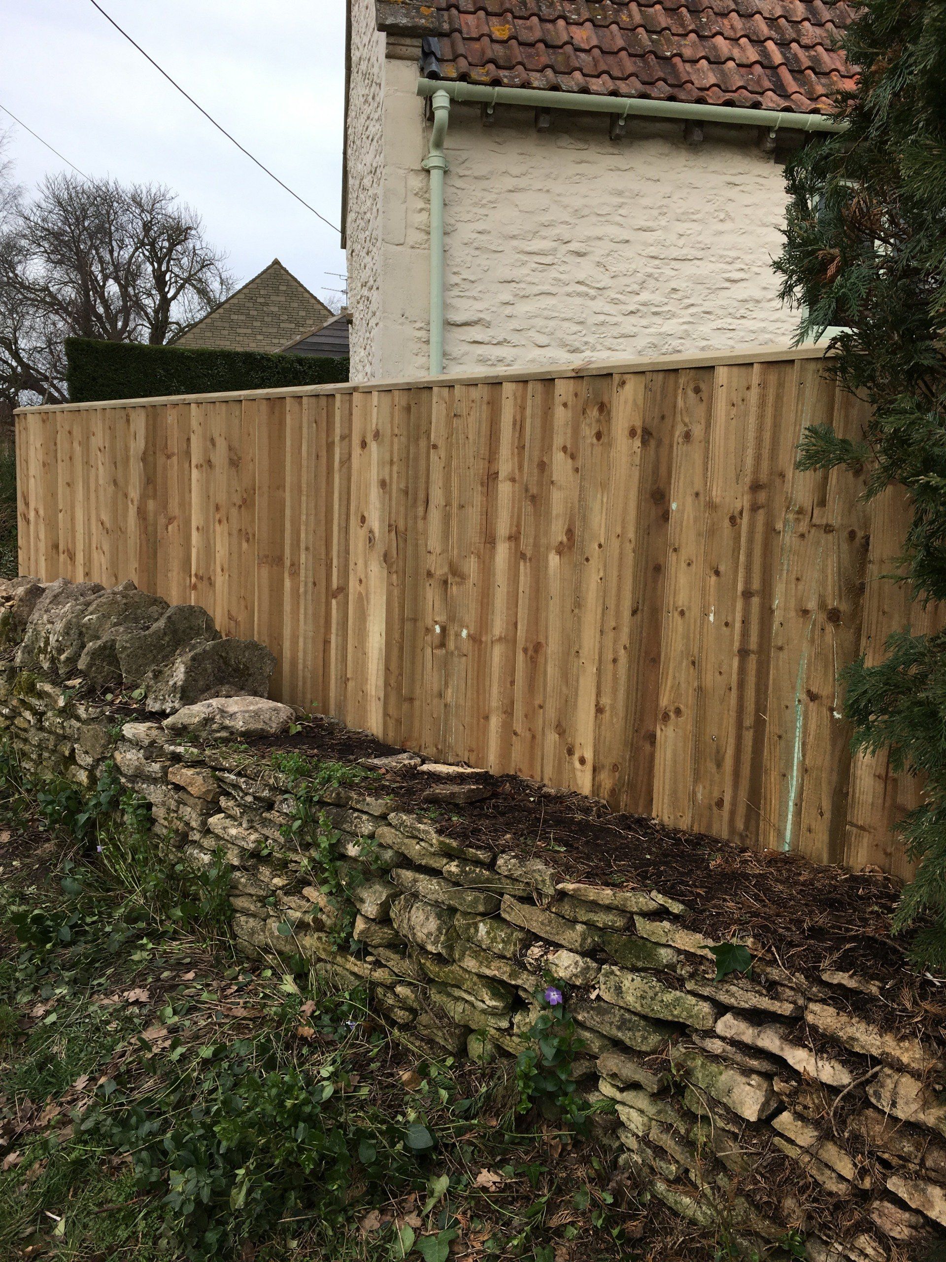 A wooden fence surrounds a stone wall next to a house.