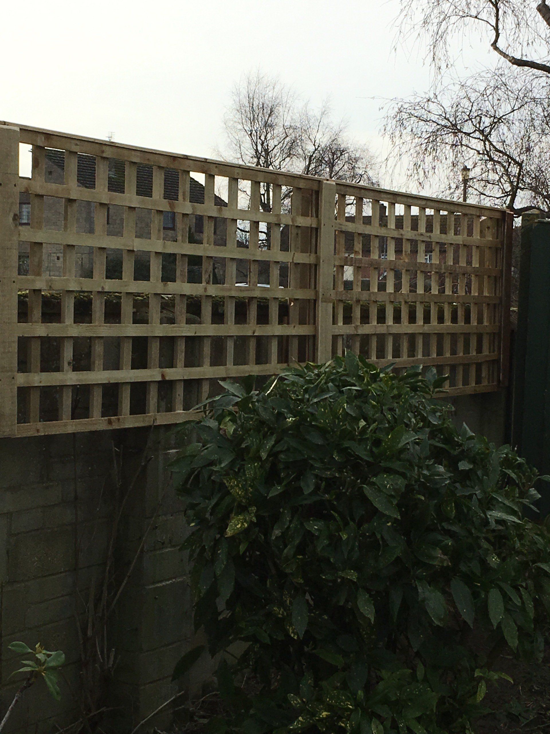 A wooden trellis is sitting on top of a concrete wall next to a bush.