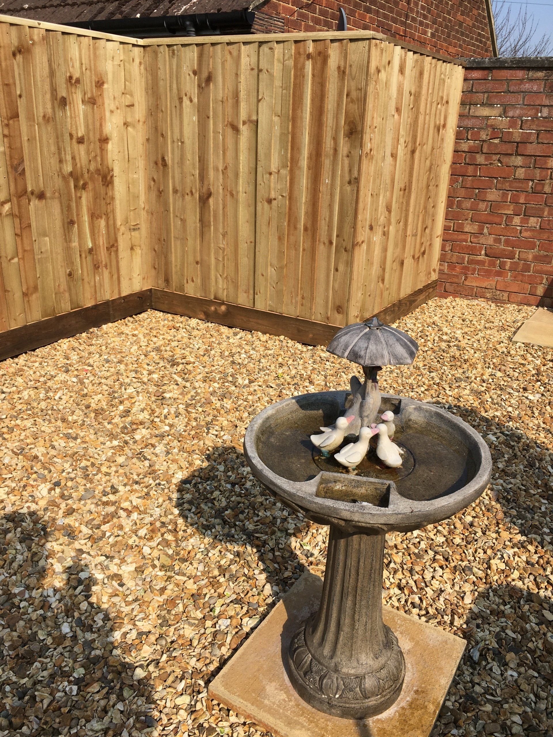 A bird bath with ducks in it is in a gravel area next to a wooden fence.