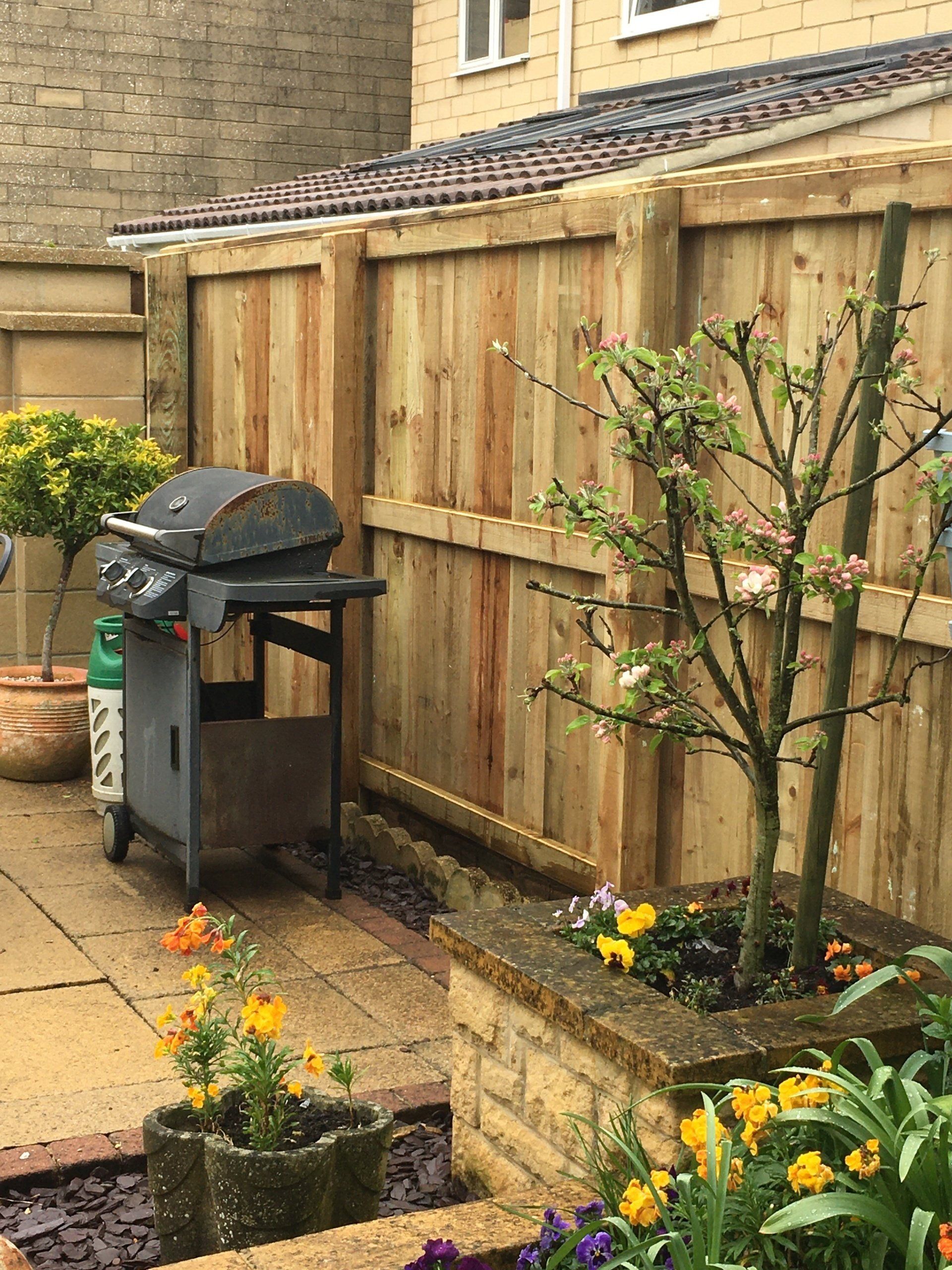A wooden fence surrounds a patio with a grill and flowers.