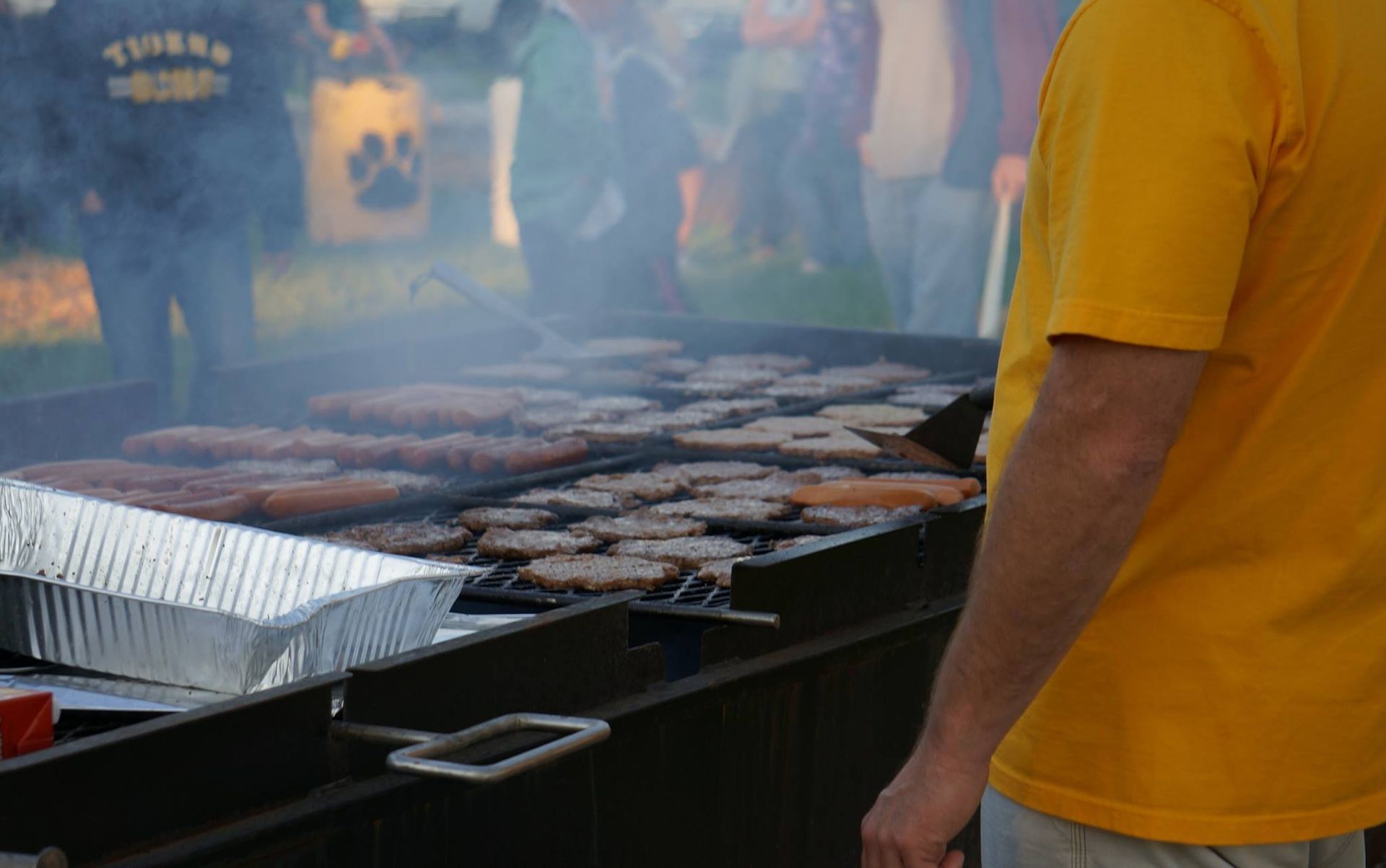 A man in a yellow shirt is standing in front of a grill cooking hot dogs.