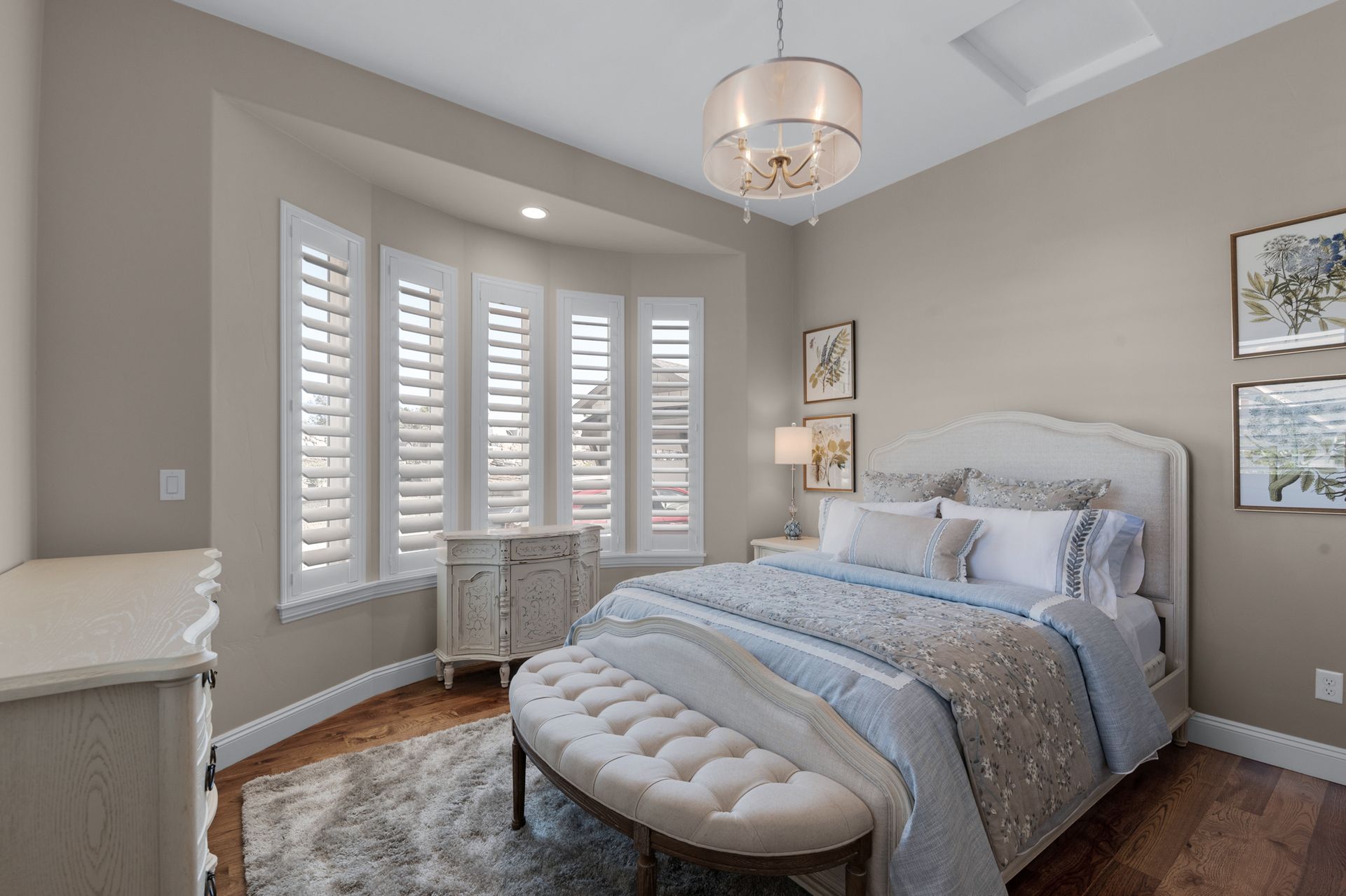 Bedroom with beige walls, bay window with shutters, bed with blue and white bedding, and a crystal chandelier.