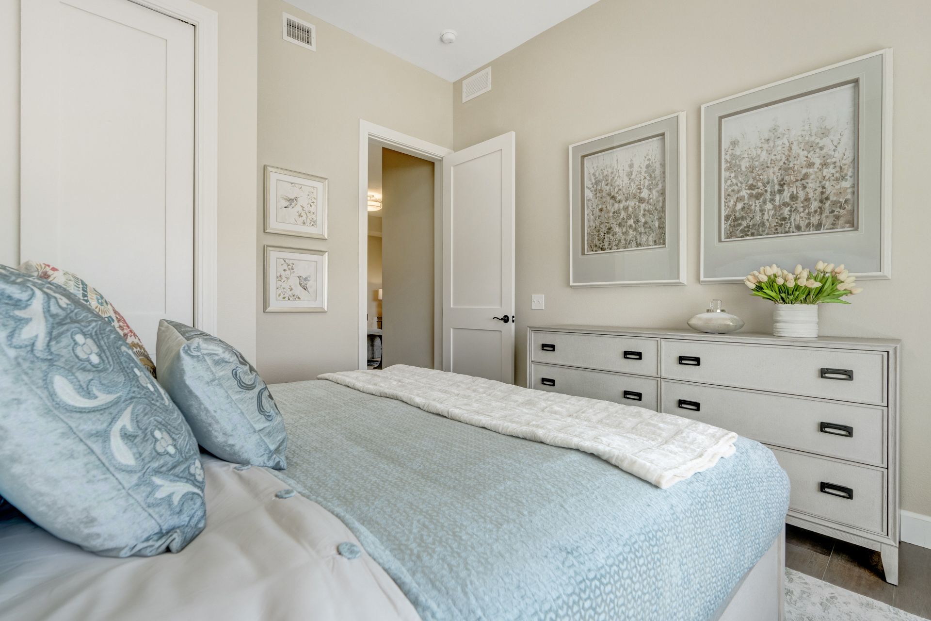 Bedroom with light blue bedding, white dresser with floral artwork, and open door.