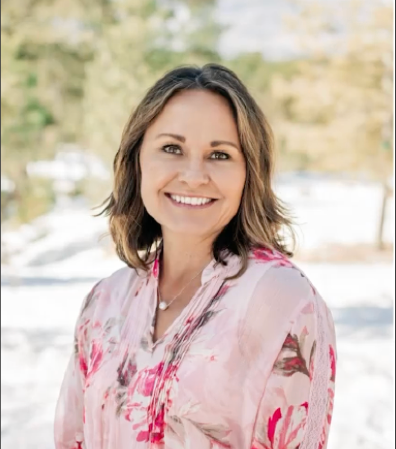 Woman with brown hair smiles, wearing a pink floral blouse, outdoors in a natural setting.