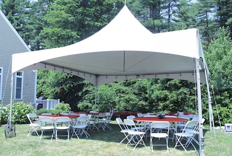 A white peaked tent set up in a backyard, sheltering round tables and folding chairs arranged for an outdoor event.