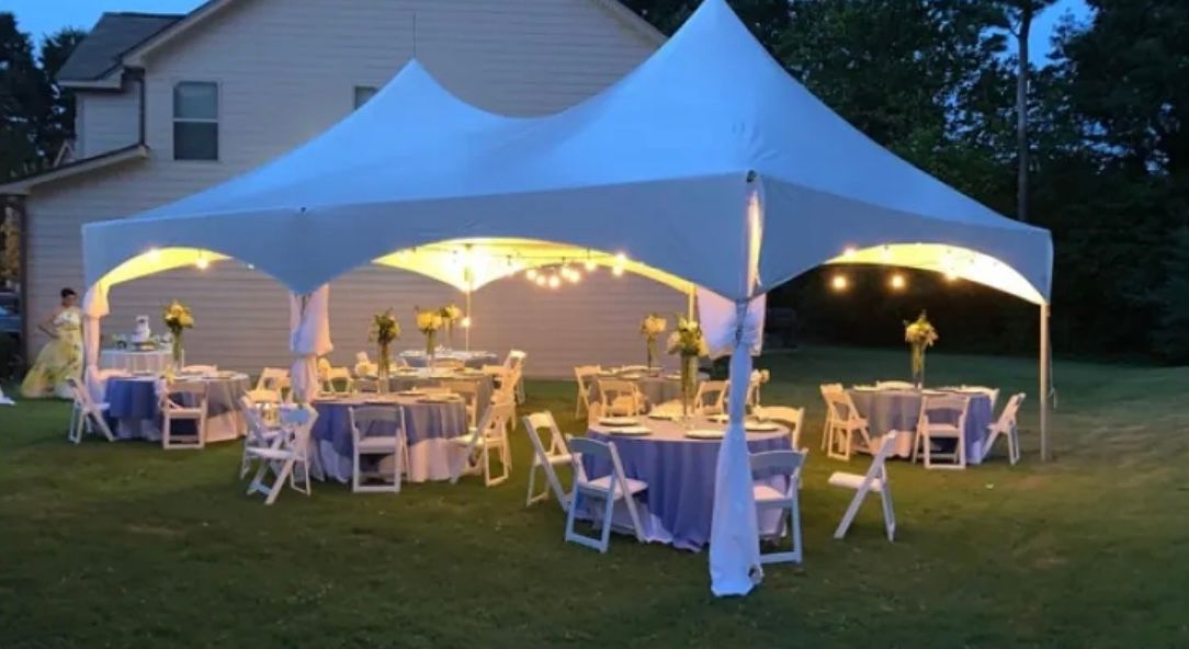 A large white event tent lit with string lights at dusk, featuring round tables with purple cloths set up on a lawn.