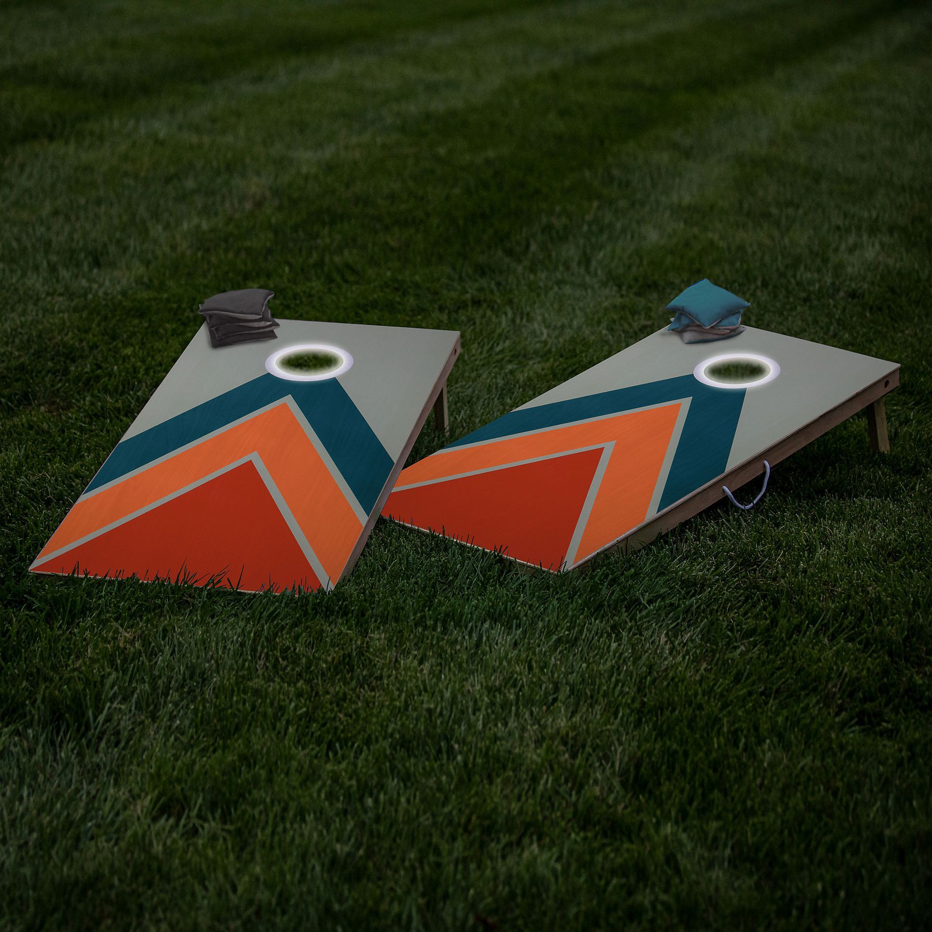 Two cornhole boards with a retro orange, teal, and grey chevron design, set up on green grass with bean bags on top.