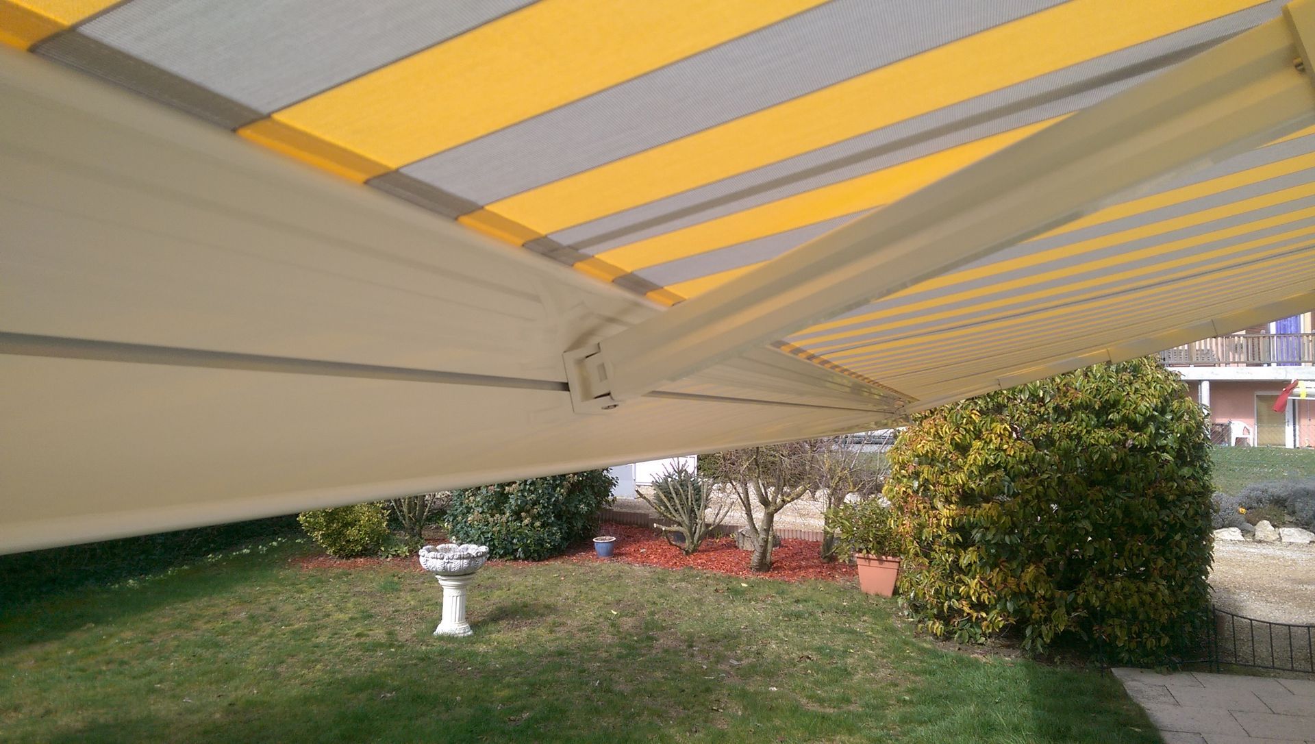 Un auvent rayé jaune et gris est suspendu au-dessus d'une cour verdoyante.