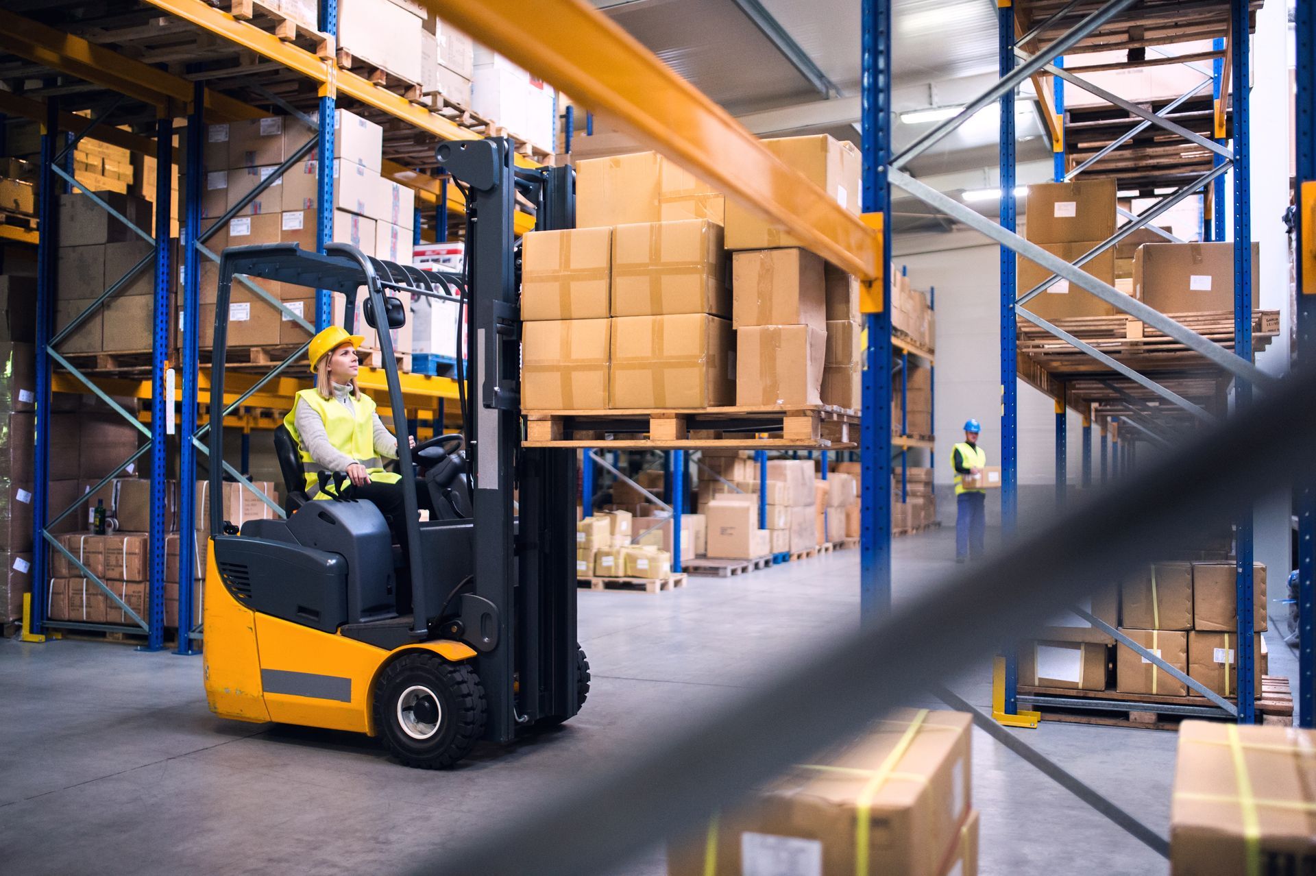 Forklift operator in a warehouse, lifting a pallet of boxes onto a high shelf.