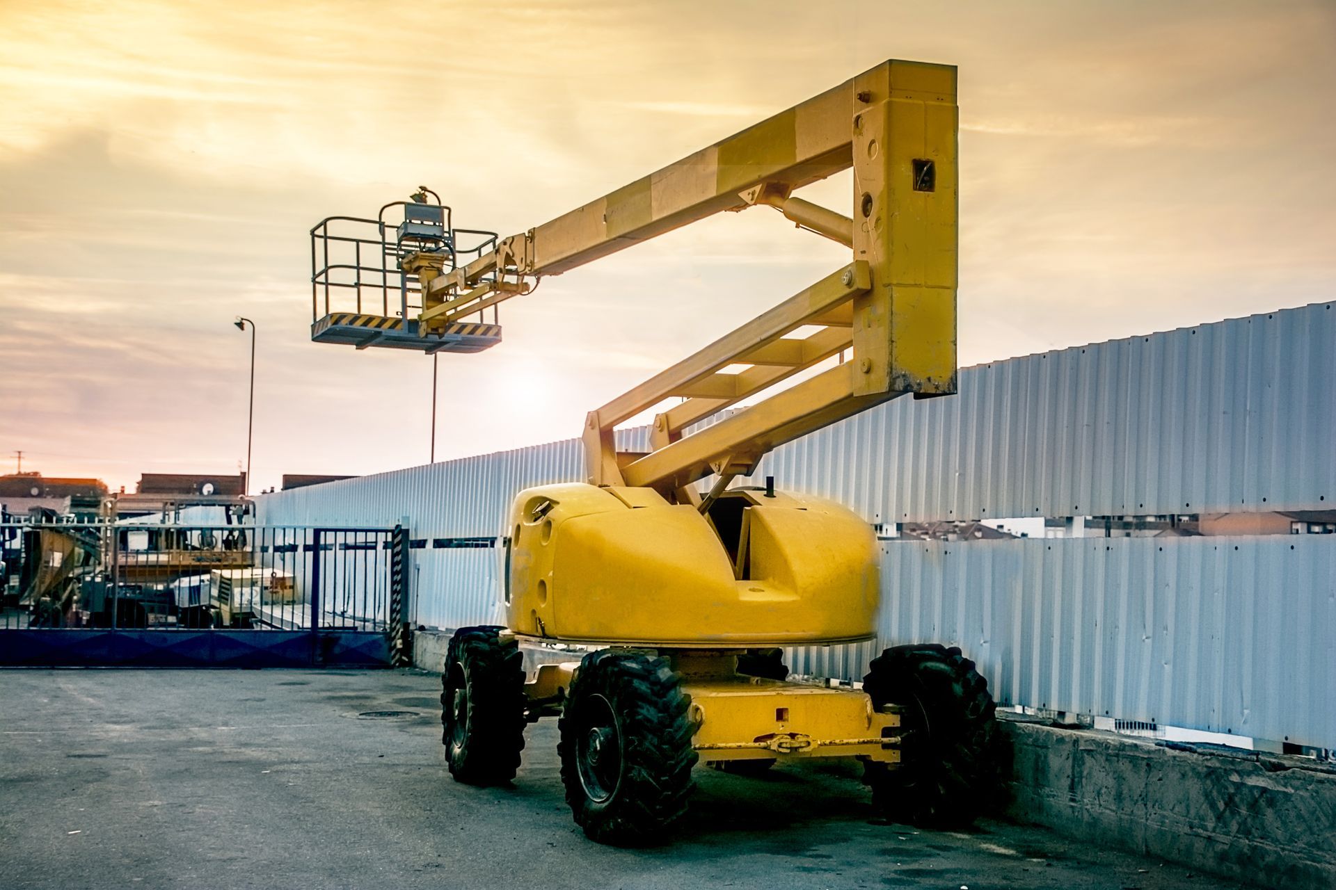 Yellow boom lift parked beside a corrugated metal fence at a worksite during sunset