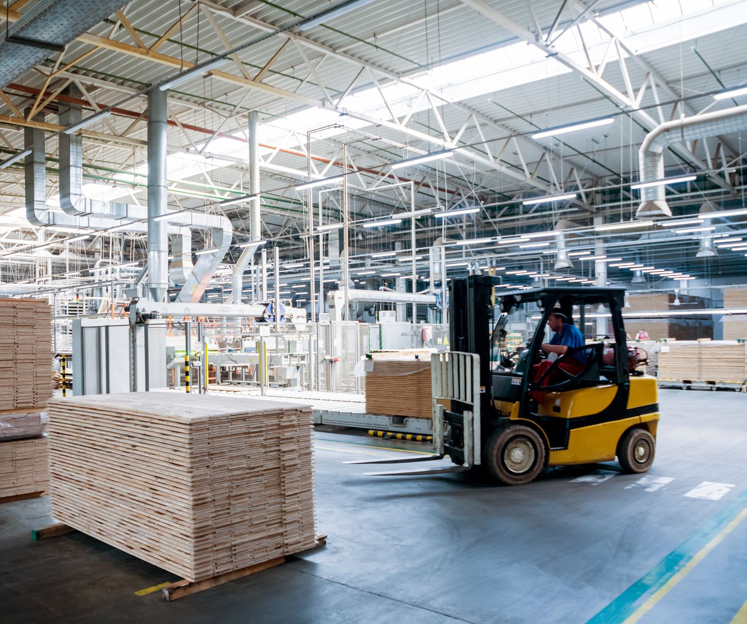 Yellow forklift in a warehouse moves stacks of lumber; a person drives.