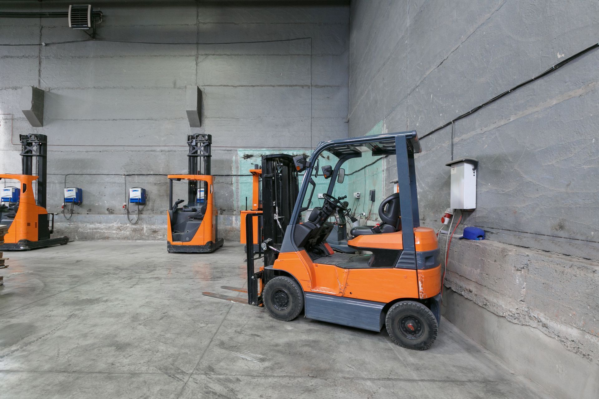 An orange forklift in a warehouse. Other forklifts are visible in the background against a concrete wall.