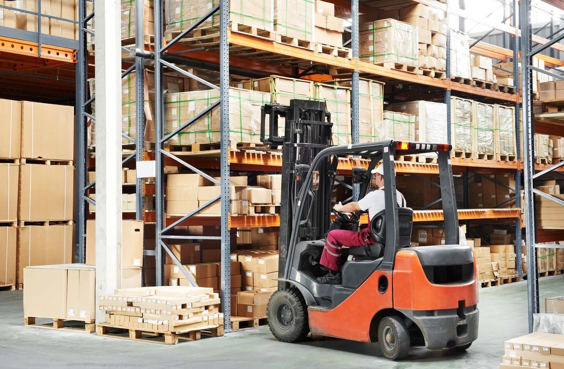 Forklift in a warehouse, moving pallets of boxes on shelves. Orange forklift, worker driving.