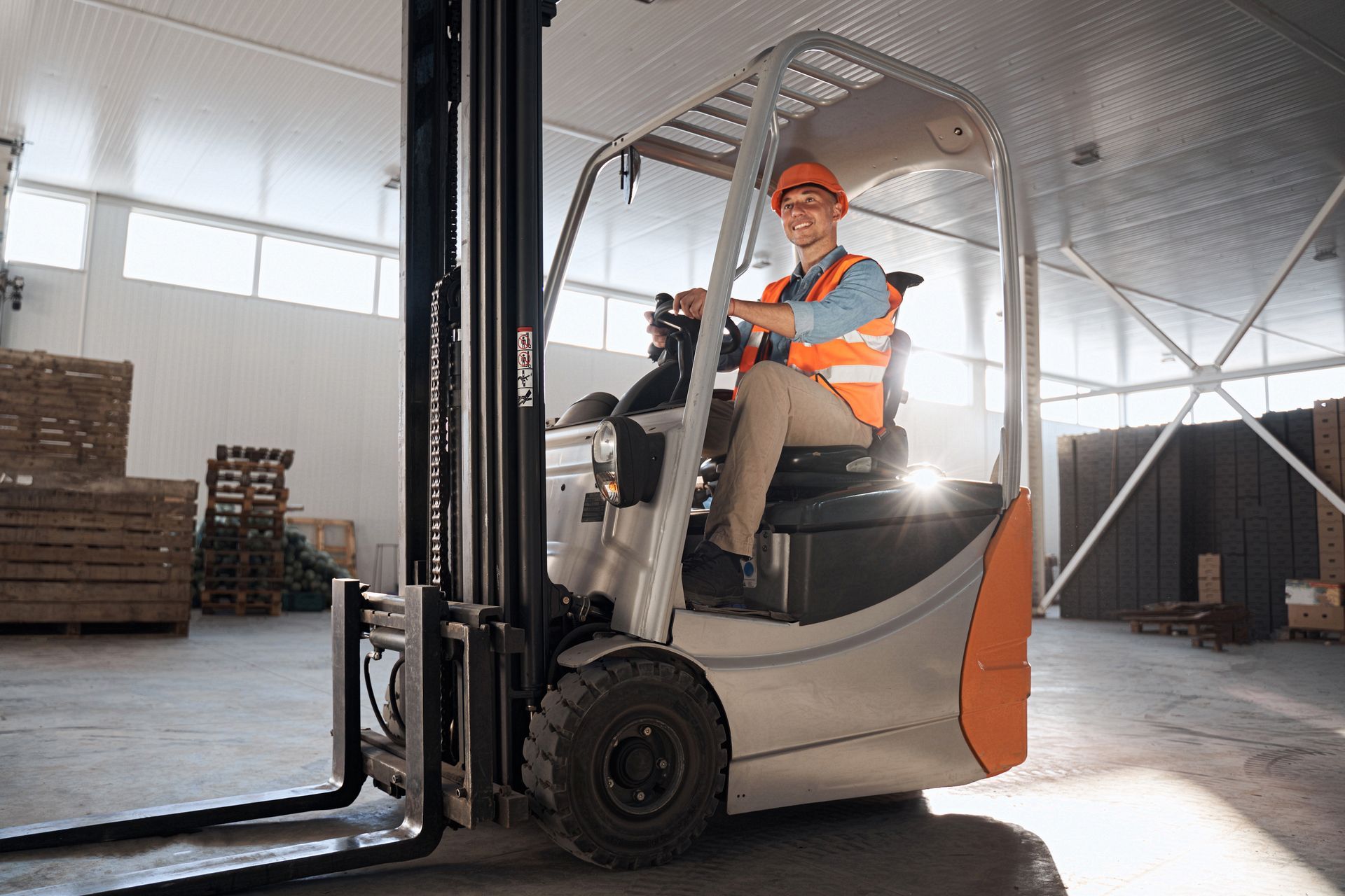 Man operating a forklift, carrying a pallet with a large cardboard box inside a warehouse.