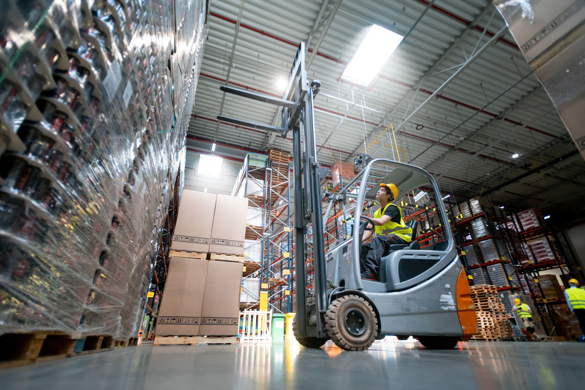 Forklift operator in a warehouse lifting a pallet of boxes. Shelves of goods in background.