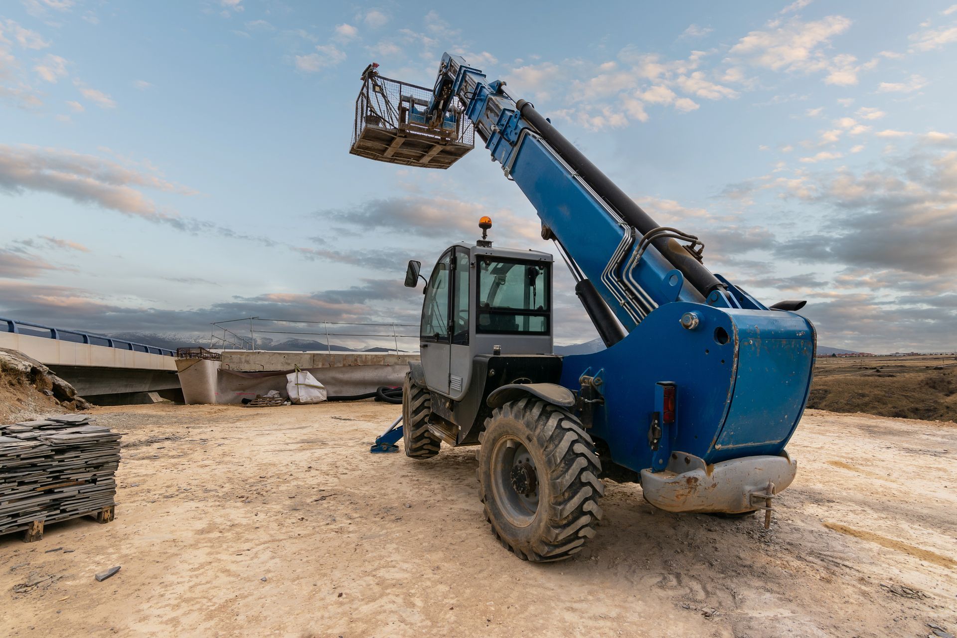 Blue telehandler on a dirt lot with raised bucket under a cloudy sky.