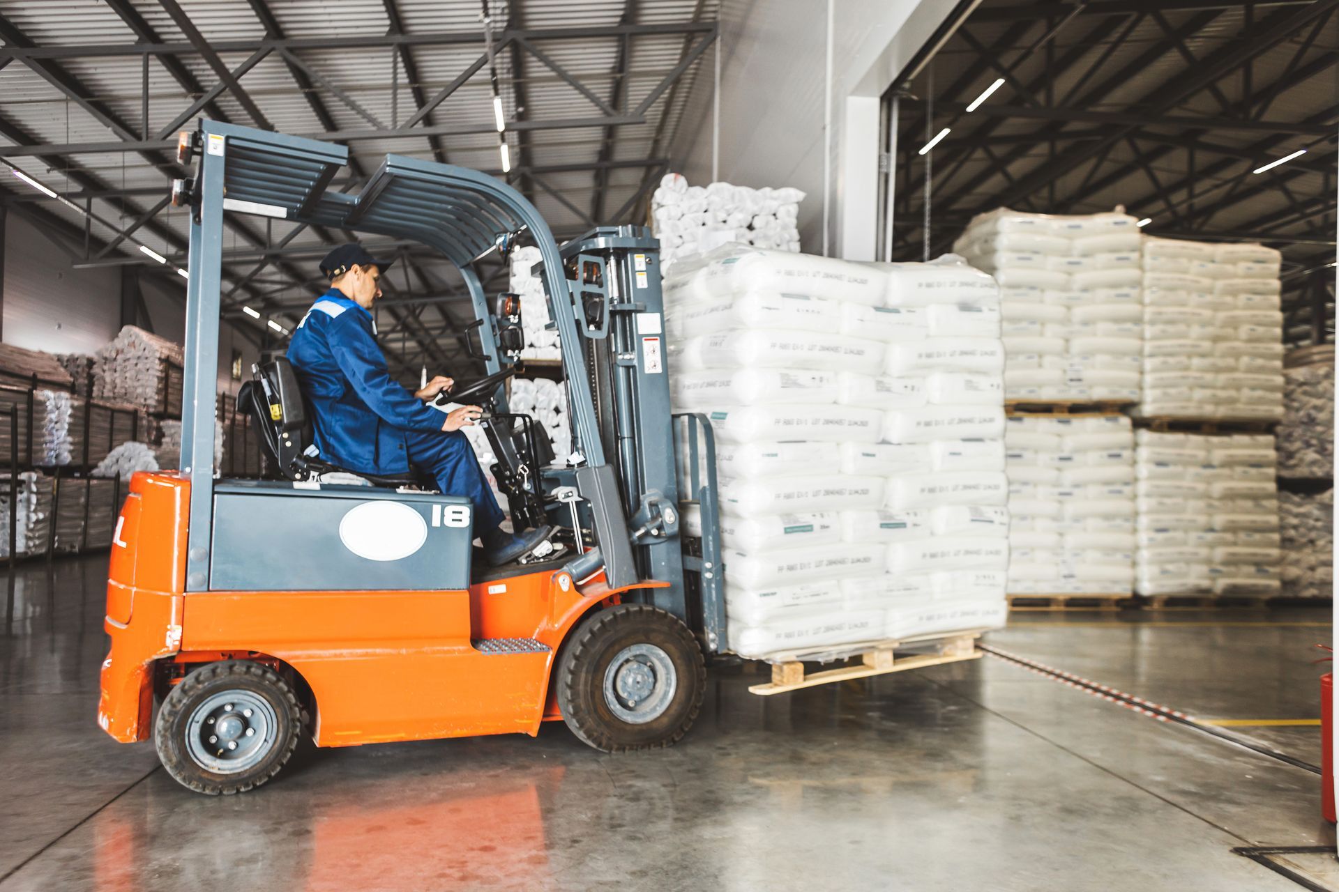 Forklift operator in a warehouse, moving a pallet of wrapped goods. Orange forklift, blue uniform.