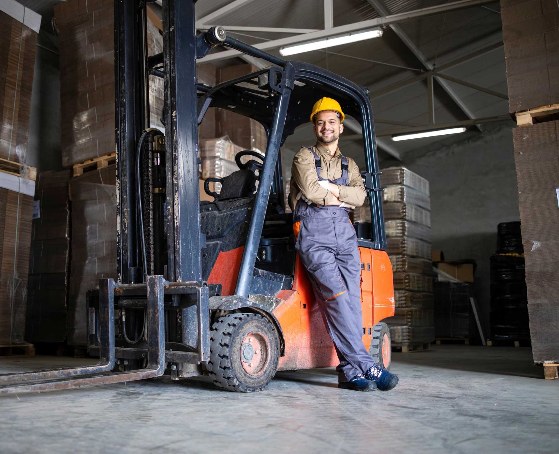 Warehouse worker leaning on forklift, smiling, wearing a hard hat, and coveralls. Inside a warehouse. Warehouse worker leaning on forklift, smiling, wearing a hard hat, and coveralls. Inside a warehouse.