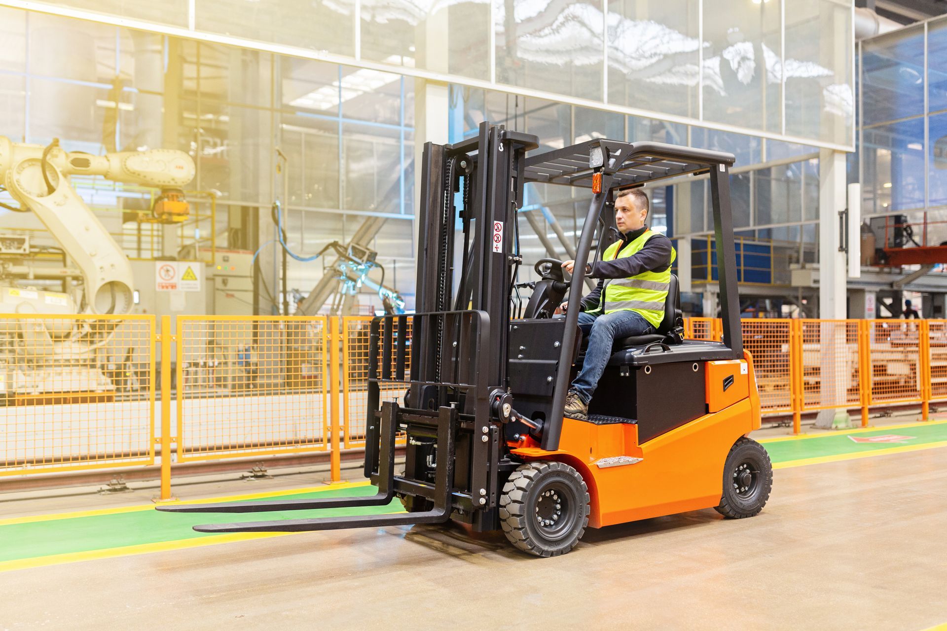Forklift operator in a factory, wearing a safety vest. Orange forklift on a factory floor with machinery.