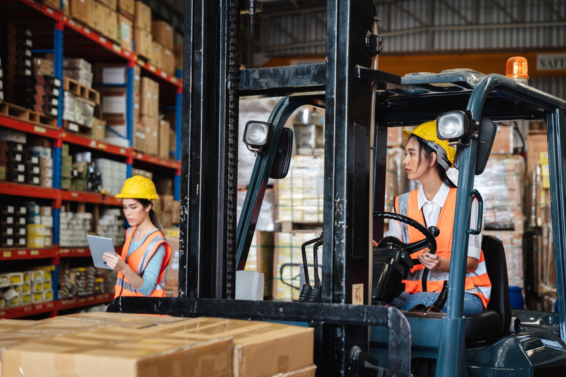 Two women in a warehouse: one driving a forklift, the other using a tablet near shelves of boxes.