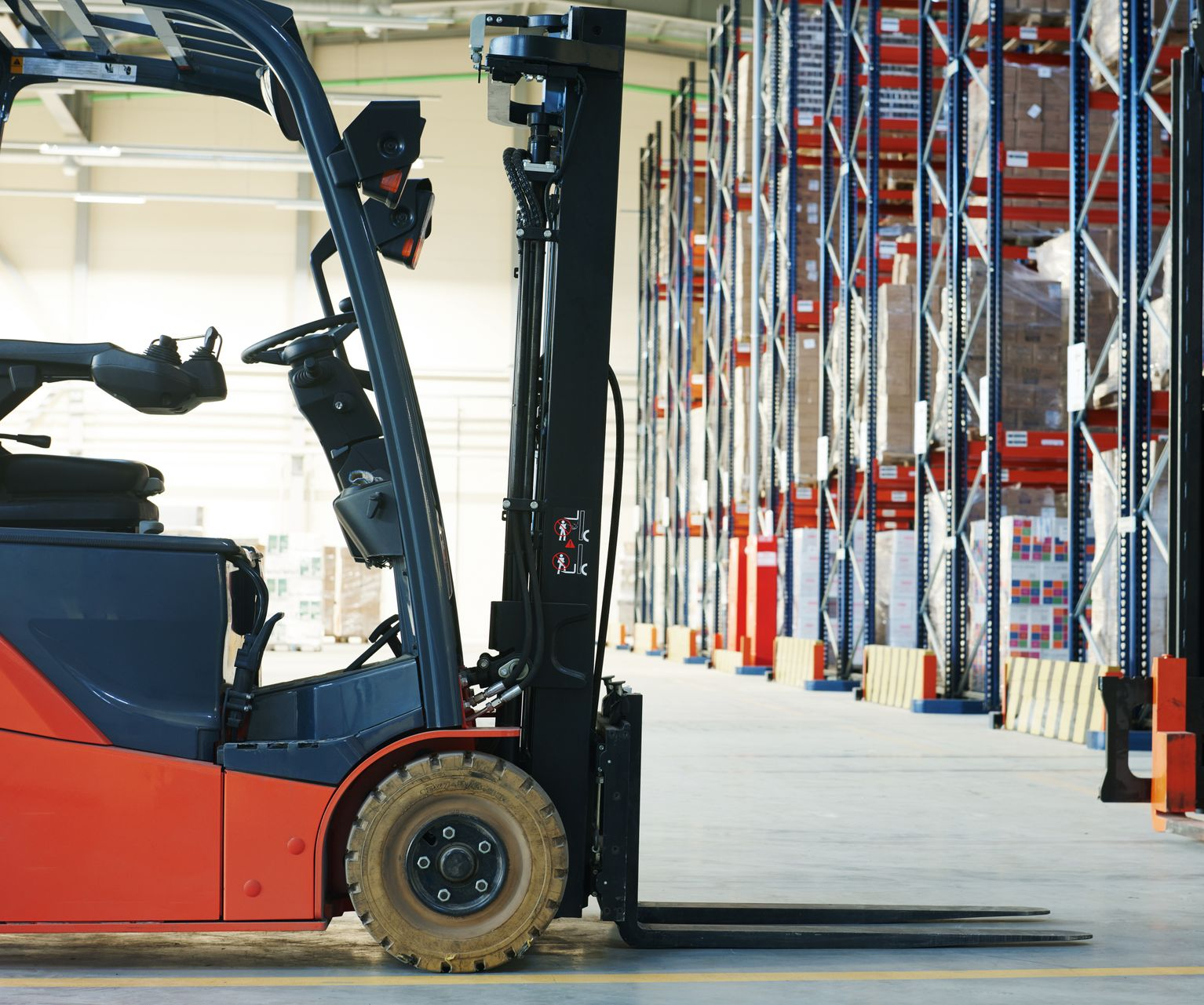 Forklift in warehouse aisle, orange and gray, with shelves of stacked boxes in the background.