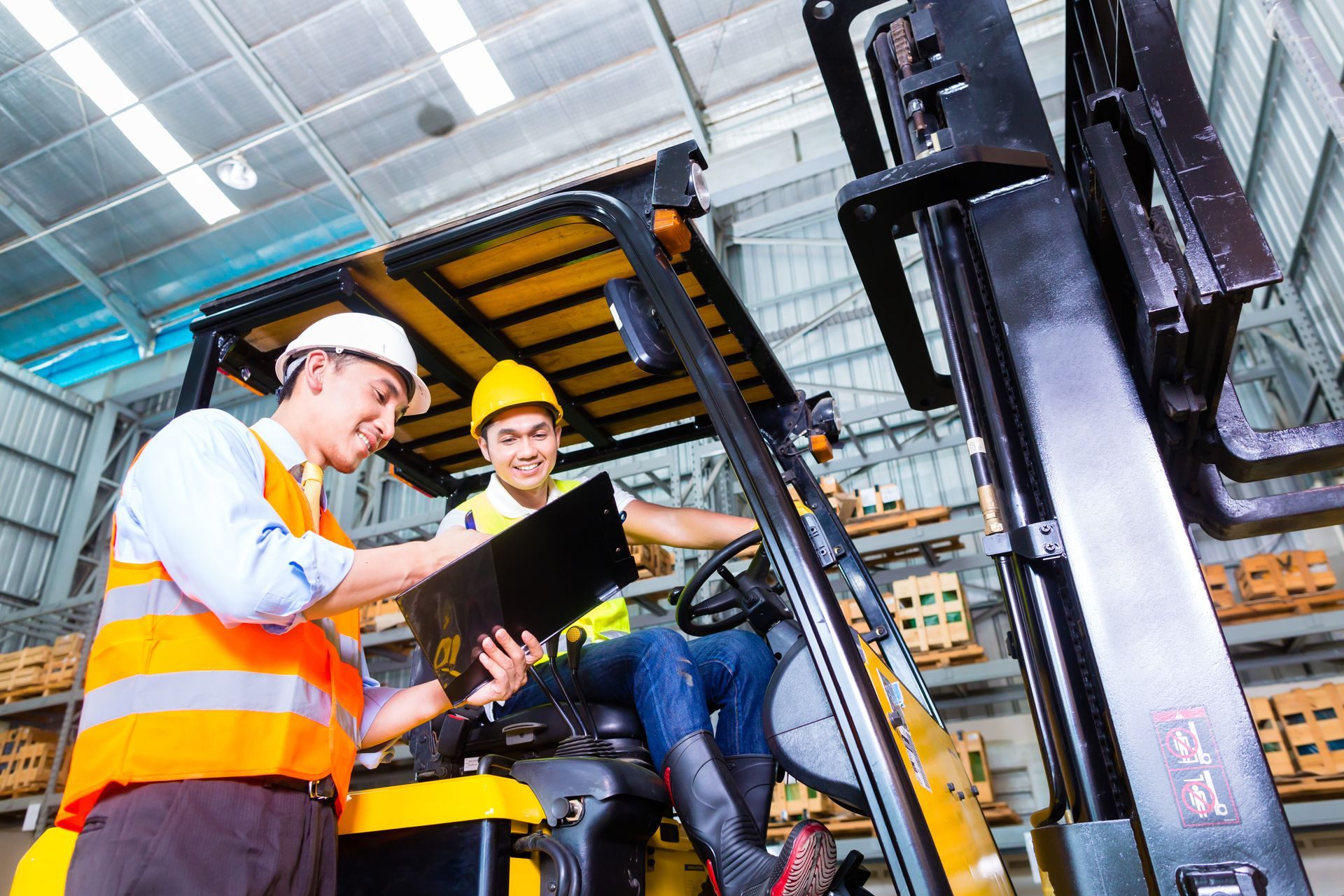 Two warehouse employees review paperwork while seated on and beside a forklift inside a warehouse.