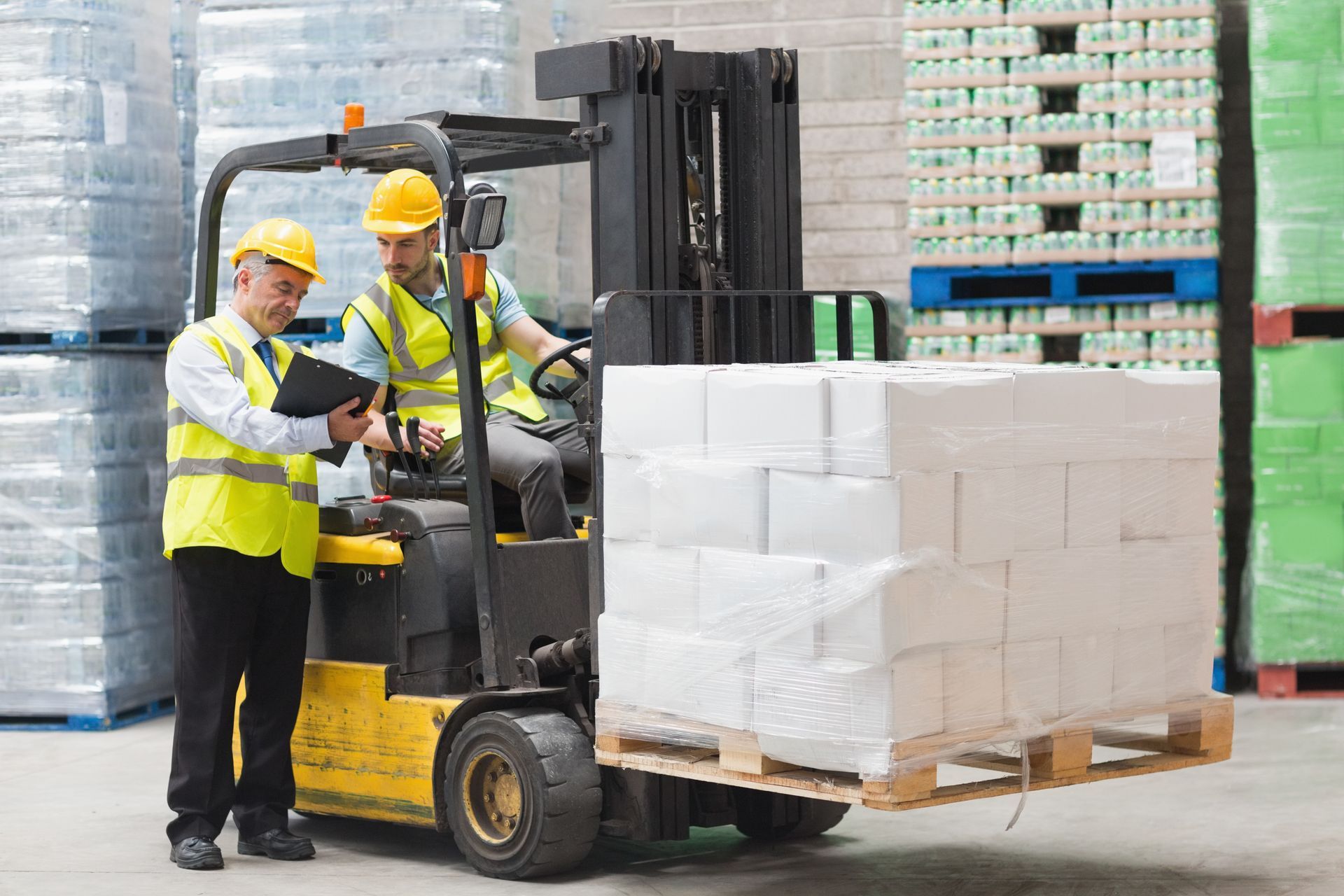 Two workers in a warehouse: One operating a forklift, the other reviewing a clipboard.